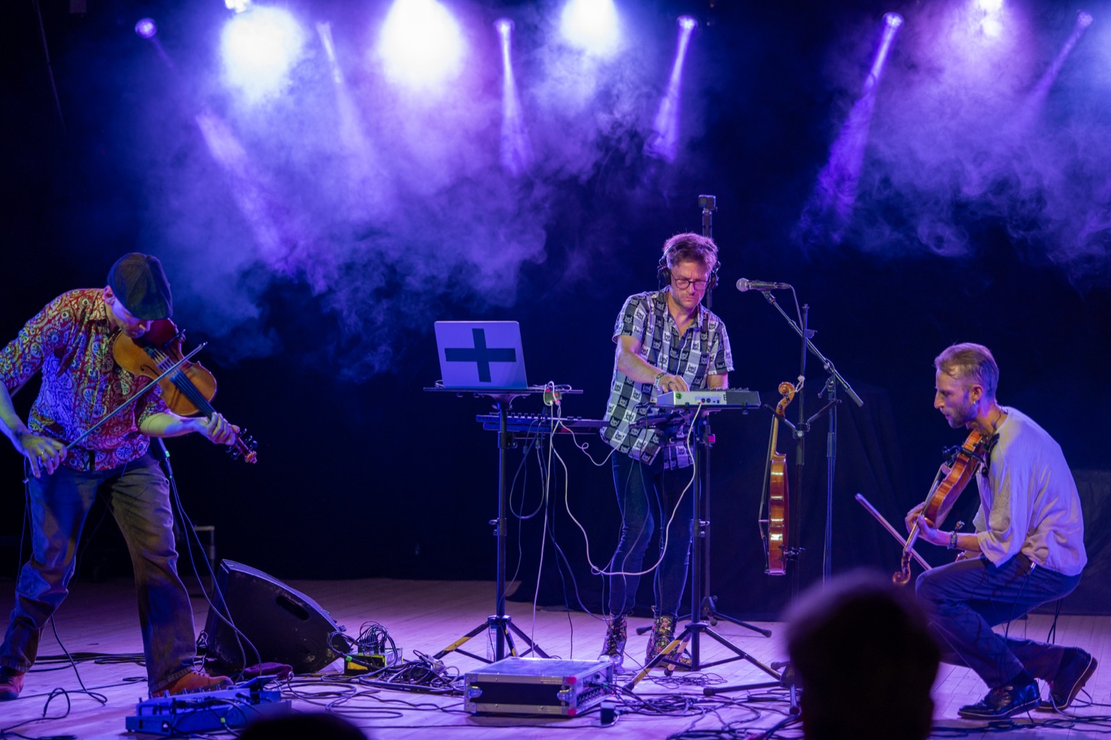 T3HO. on stage in violet haze: one fiddler on the left, Antti Puumalainen at the electronics in the middle, the other fiddler kneeling on the right